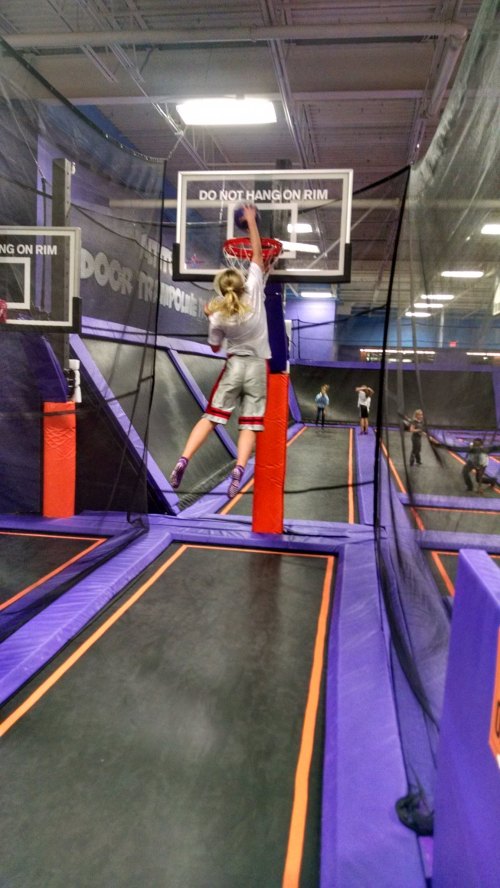 A child jumps to dunk a basketball at a trampoline park, surrounded by safety nets and other jumpers.