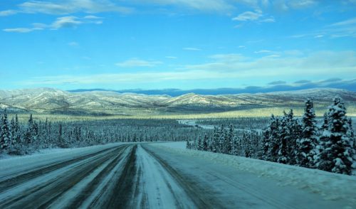 A snowy road winds through a winter landscape of mountains and evergreen trees under a blue sky.