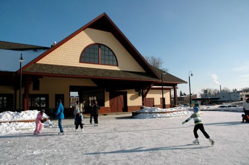 Children ice skating on a frozen surface near a large building, with snow and clear blue skies in the background.