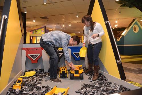 A man and woman play with toy construction trucks in a children's activity area filled with black stones.