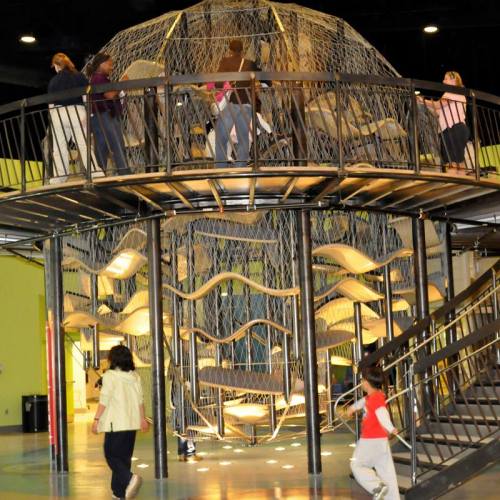 A multi-level play structure with a netted design, featuring children and adults exploring and climbing.