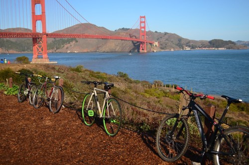 Bicycles parked along a path with the Golden Gate Bridge and bay in the background on a clear day.