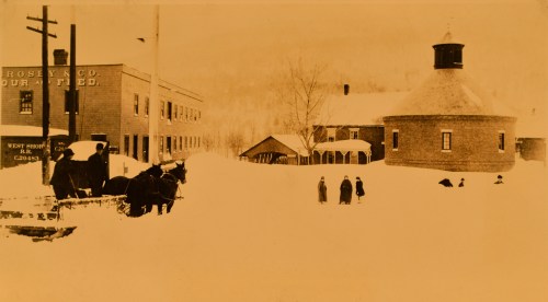 Historic winter scene with snow-covered buildings, horses, and people in a rural setting.