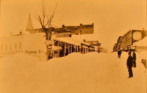Historic snowy street scene with buildings and signs partially buried in snow, two figures standing in the foreground.