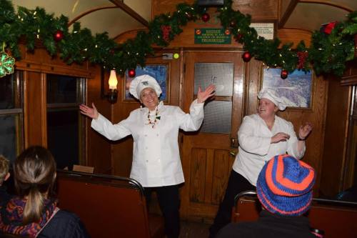 Two women in chef hats perform joyfully inside a decorated train car, with festive garlands and an audience watching.