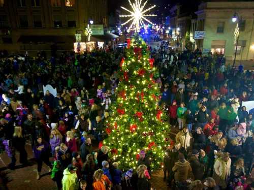 A large crowd gathers around a beautifully lit Christmas tree in a festive downtown setting at night.