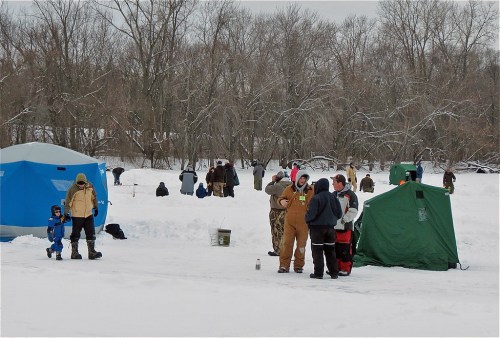 People ice fishing on a snowy lake, with tents and equipment set up in a winter landscape.
