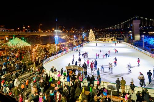 A bustling outdoor ice skating rink at night, surrounded by festive lights and a large Christmas tree.