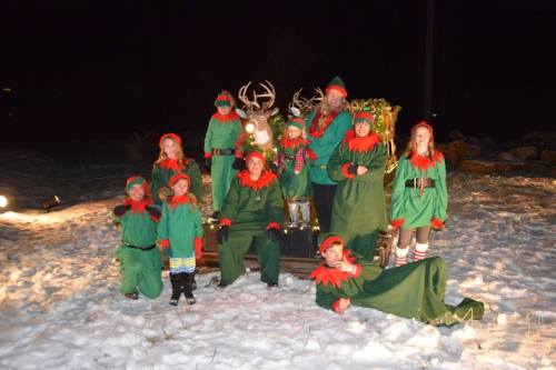 A group of people dressed as elves poses in the snow, with festive decorations and reindeer in the background.
