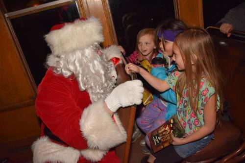 Santa Claus interacts with three young girls on a festive train, sharing smiles and holiday cheer.