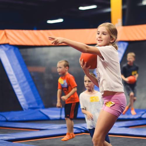A girl smiles while playing with a basketball on a trampoline, surrounded by other children in a colorful indoor space.