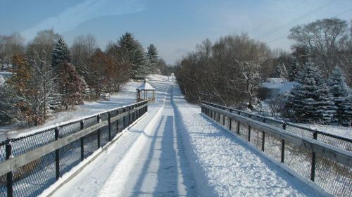 A snowy path leads through a winter landscape, with trees and a gazebo in the distance under a clear blue sky.