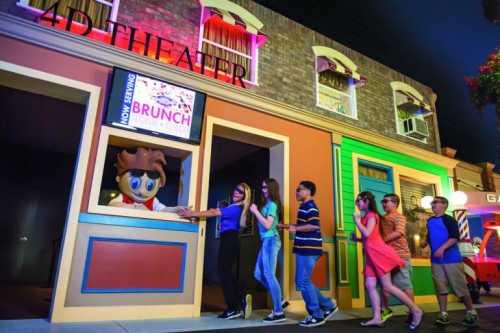 A group of children and adults line up at a colorful theater entrance with a mascot serving brunch.