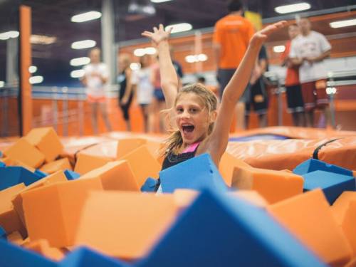 A joyful girl with arms raised sits in a colorful foam pit at a trampoline park, surrounded by other kids.