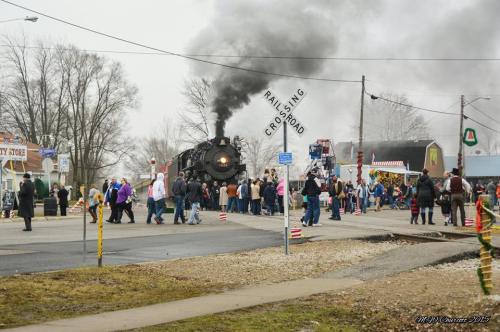 A steam train billows smoke as a crowd gathers near a railroad crossing, with shops and trees in the background.