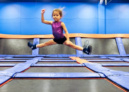 A young girl in a purple top jumps joyfully on a trampoline, with her legs spread wide in mid-air.