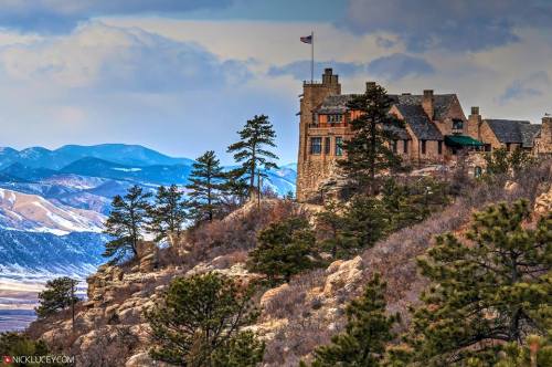 A stone castle perched on a rocky hill, surrounded by pine trees and mountains under a cloudy sky.