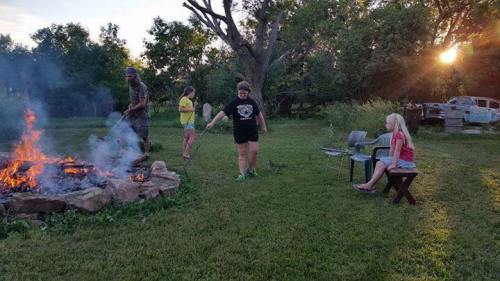 A group of people gathered around a campfire in a grassy area, with trees and an old vehicle in the background.