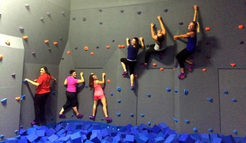 A group of six people climbing a colorful indoor rock wall, with foam blocks below for safety.
