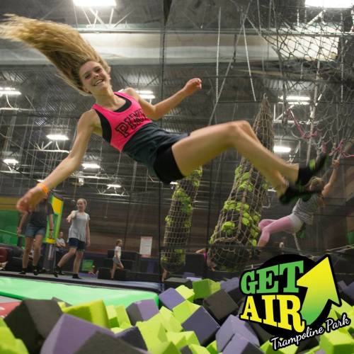 A girl jumps on a trampoline, surrounded by colorful foam blocks, with others playing in the background at a trampoline park.