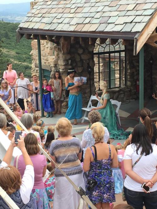 A crowd watches a performer in a blue costume reading to children in a scenic outdoor setting.