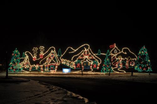 Colorful holiday lights outline a festive scene with trees, mountains, and whimsical decorations against a night sky.
