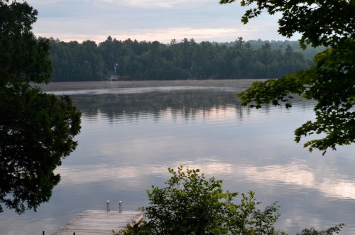 A calm lake reflects the sky, surrounded by trees, with a wooden dock extending into the water.