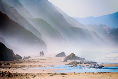A couple walks along a misty beach, with soft hills and rocky formations in the background.
