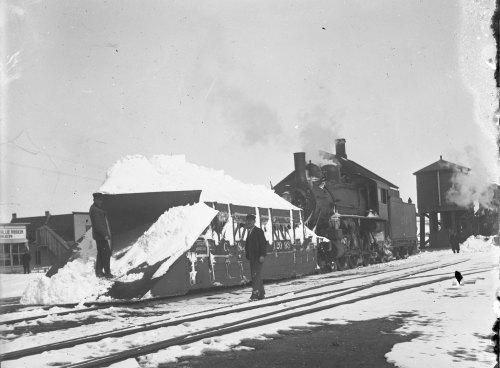 A vintage steam locomotive with a snowplow clears snow from train tracks in a snowy landscape.