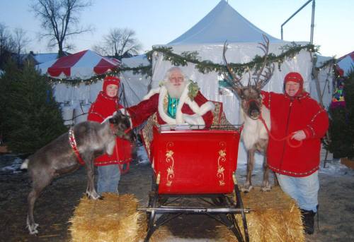 Santa Claus sits in a red sleigh surrounded by two reindeer and two people in red coats, with festive decorations in the background.