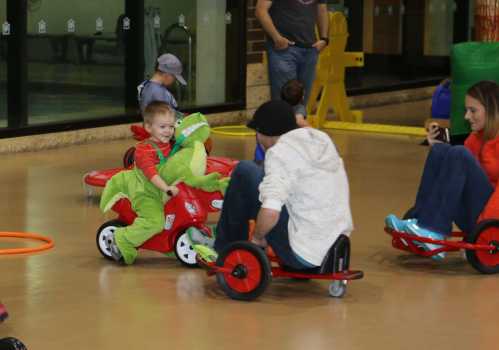 Children playing in a gym, riding tricycles; one child wears a dinosaur costume while another sits on a tricycle.