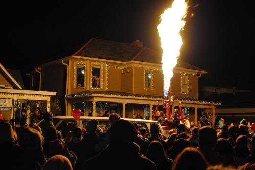 A crowd gathers at night as a flame shoots up from a vehicle, with a decorated house in the background.