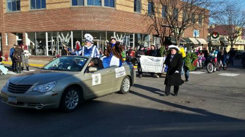 A parade scene with a convertible car, people in costumes, and a crowd watching along the street.