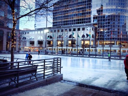 A city ice skating rink with people skating, surrounded by tall buildings and festive lights.