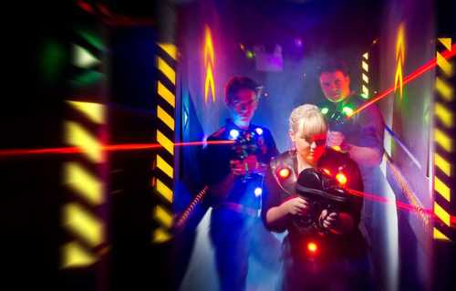 Three people in a dark, colorful laser tag arena, with bright lights and smoke creating a dynamic atmosphere.