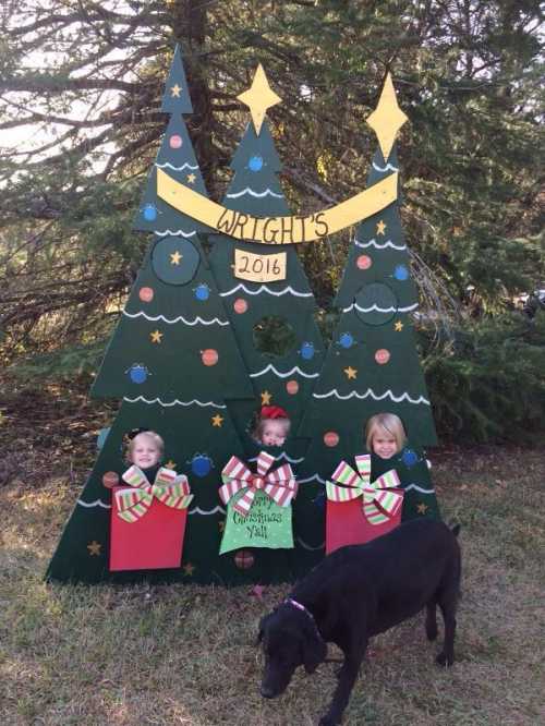 Three children peek out from a festive Christmas tree display, holding large gift boxes, with a dog nearby.