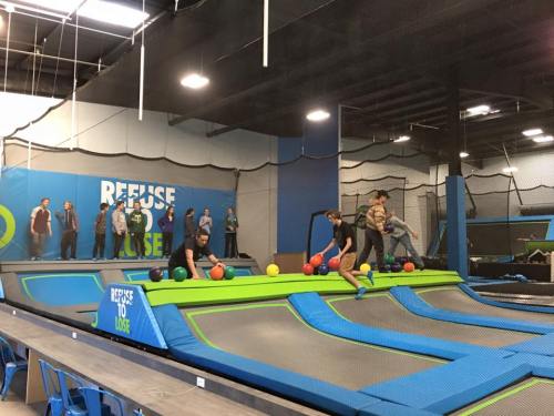 A trampoline park scene with people bouncing and playing dodgeball on trampolines, while others watch from the sidelines.