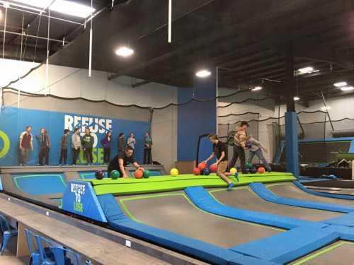 A trampoline park scene with people bouncing and playing dodgeball on trampolines, while others watch from the sidelines.