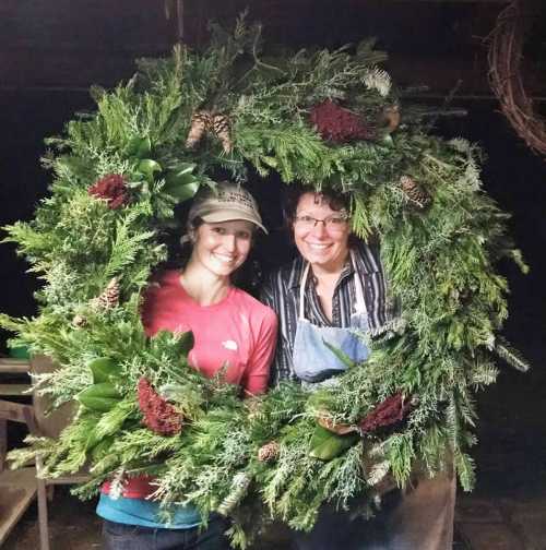 Two smiling individuals stand inside a large, lush green wreath adorned with pinecones and red accents.