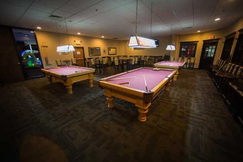 A spacious game room featuring three pool tables with purple felt and overhead lights, surrounded by seating.