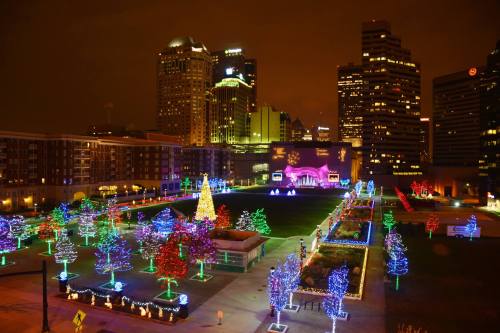 A city park decorated with colorful holiday lights, featuring trees and a large illuminated display against a skyline backdrop.