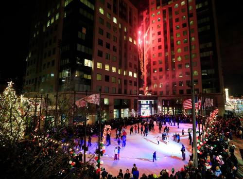 A festive ice skating rink surrounded by a crowd, with fireworks lighting up the night sky above a tall building.