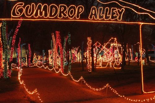 A festive pathway lit with colorful lights, marked by a sign reading "Gumdrop Alley," surrounded by illuminated trees.