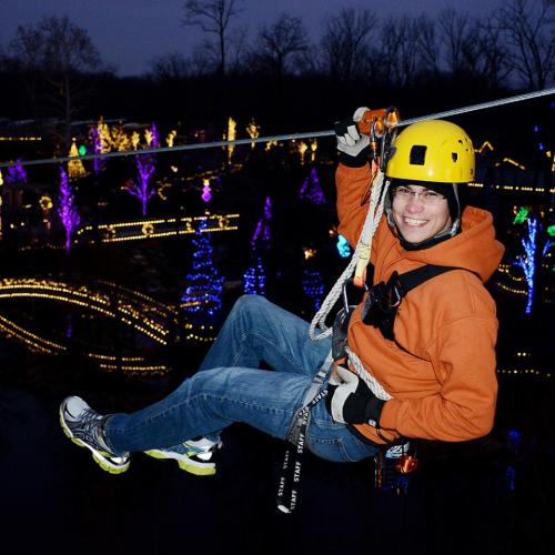 A person in a helmet and orange jacket zip-lining over a colorful, illuminated landscape at night.