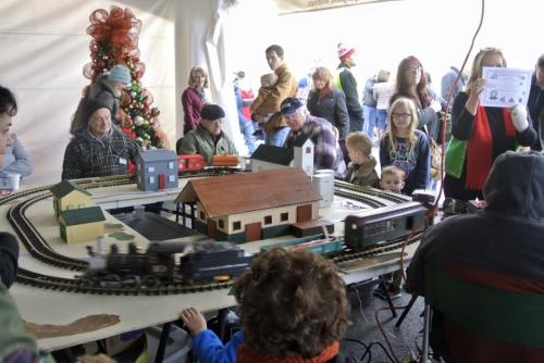 A model train setup with people watching, surrounded by festive decorations and children enjoying the display.