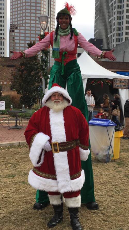 A tall performer in festive attire stands on stilts above a man in a Santa costume at a holiday event.