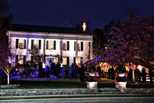 A historic house illuminated at night, with people gathered outside, festive decorations, and a warm, inviting atmosphere.