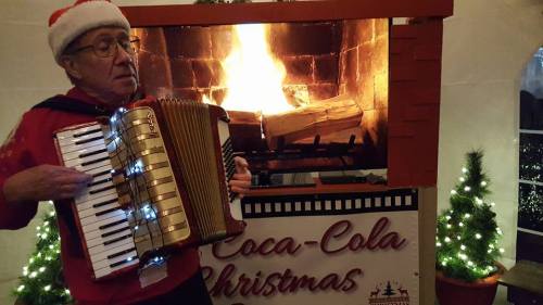 A man in a Santa hat plays an accordion in front of a festive fireplace with Christmas decorations.