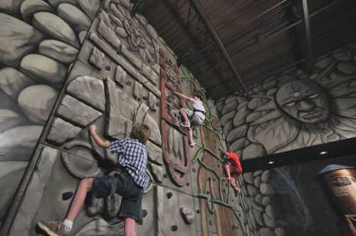 Two children climbing a textured indoor rock wall with a mural of faces and flowers in the background.