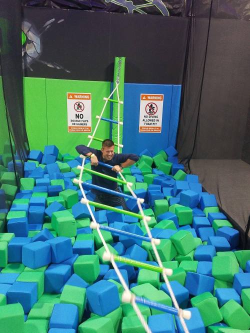 A person climbs a rope ladder above a pit filled with blue and green foam blocks in a trampoline park.